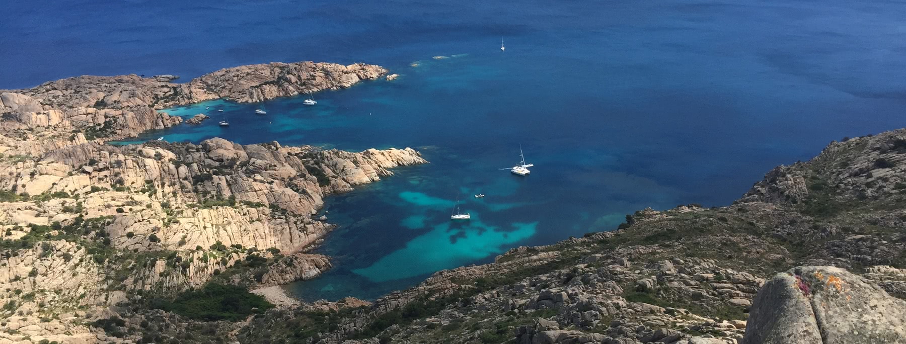 Blick auf eine kleine Bucht mit klarem, blauem Wasser, kleinen Segelbooten und einer felsigen Küste